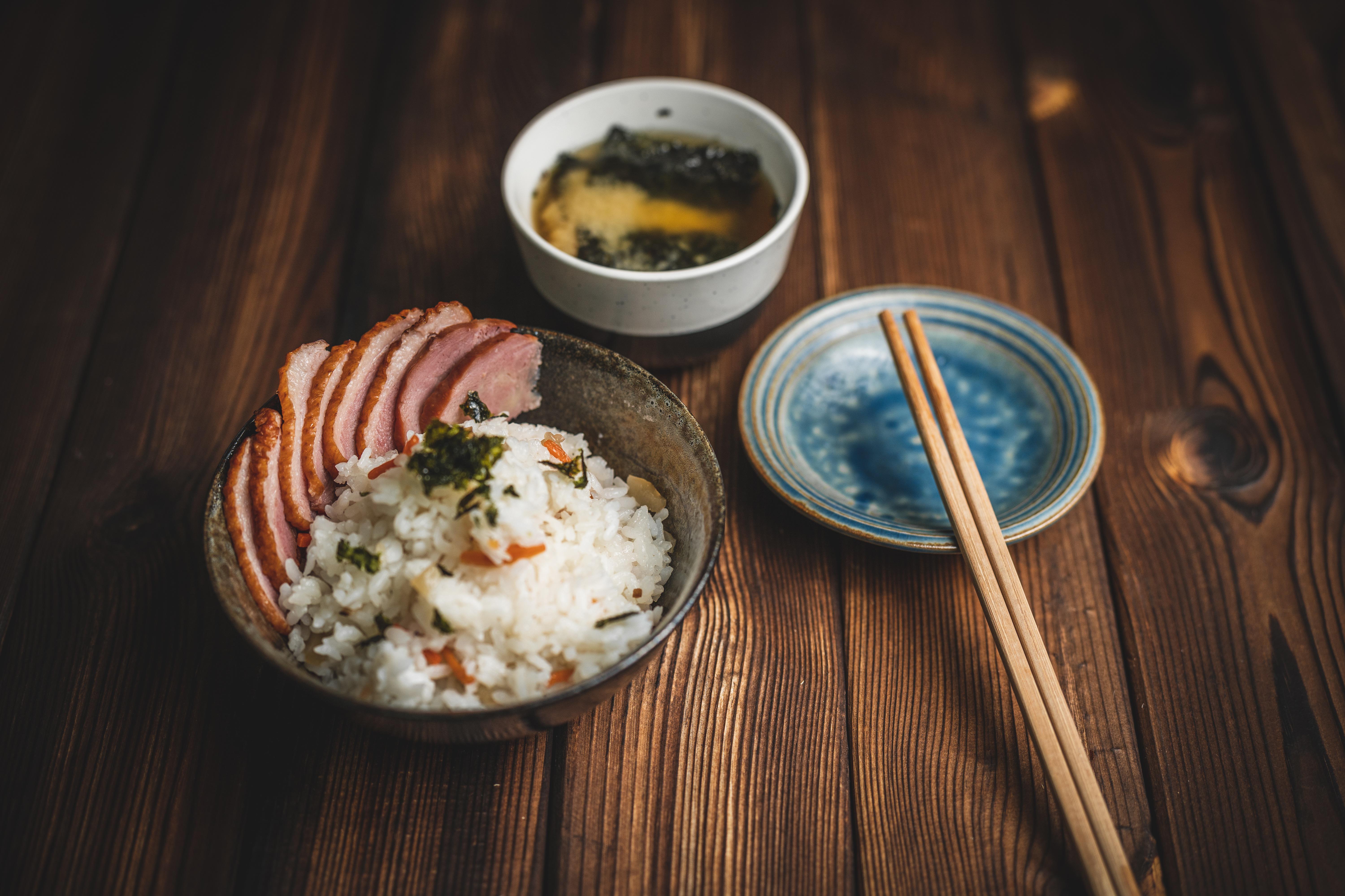 A bowl of rice topped with sliced smoked duck and nori, beside a bowl of soup and chopsticks resting on dark wood