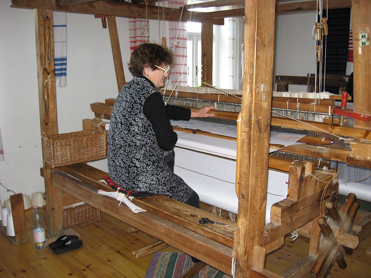 A woman seated at a large wooden hand loom, working the warp threads